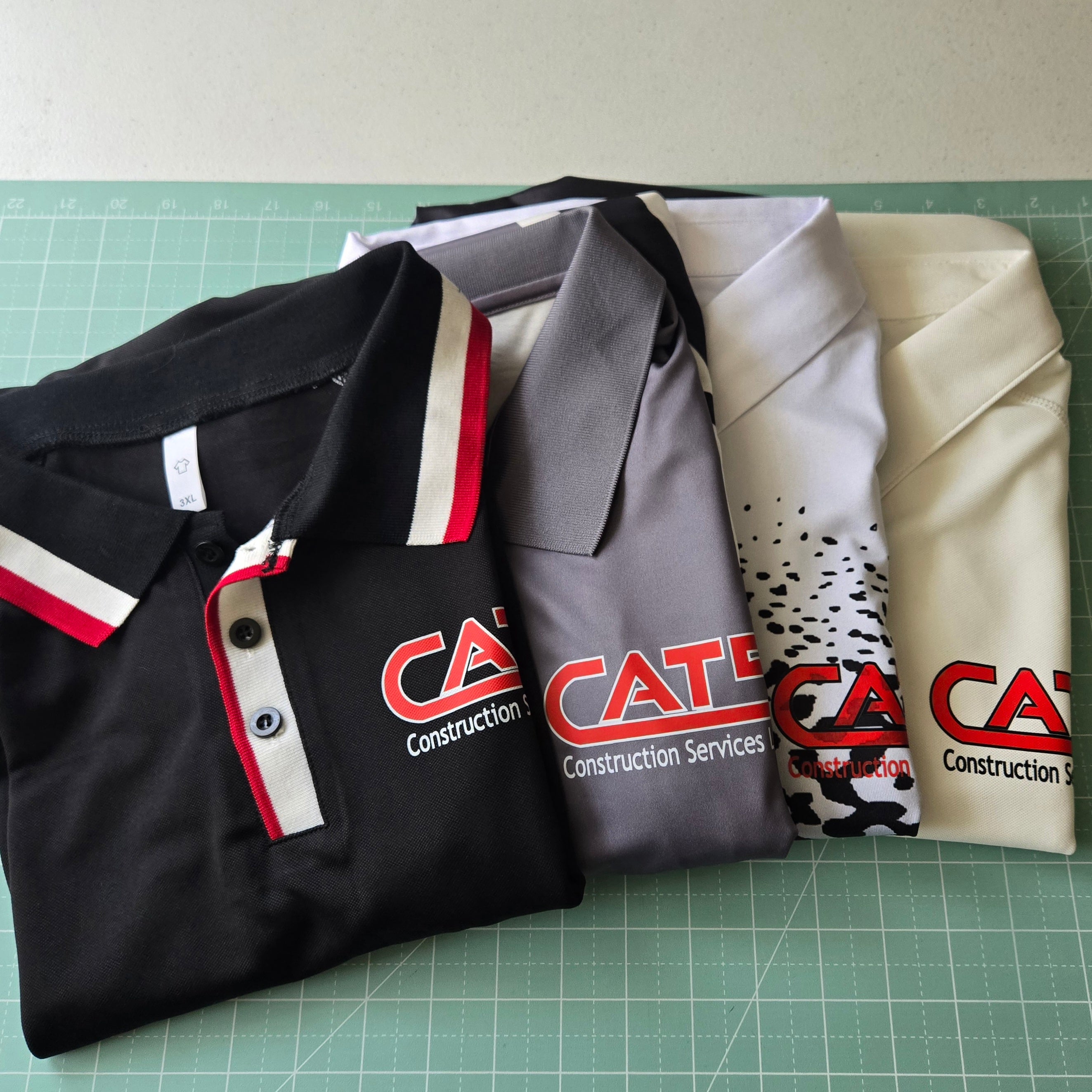 Four polo shirts with 'Caterpillar Construction Services' logos on a green cutting mat.