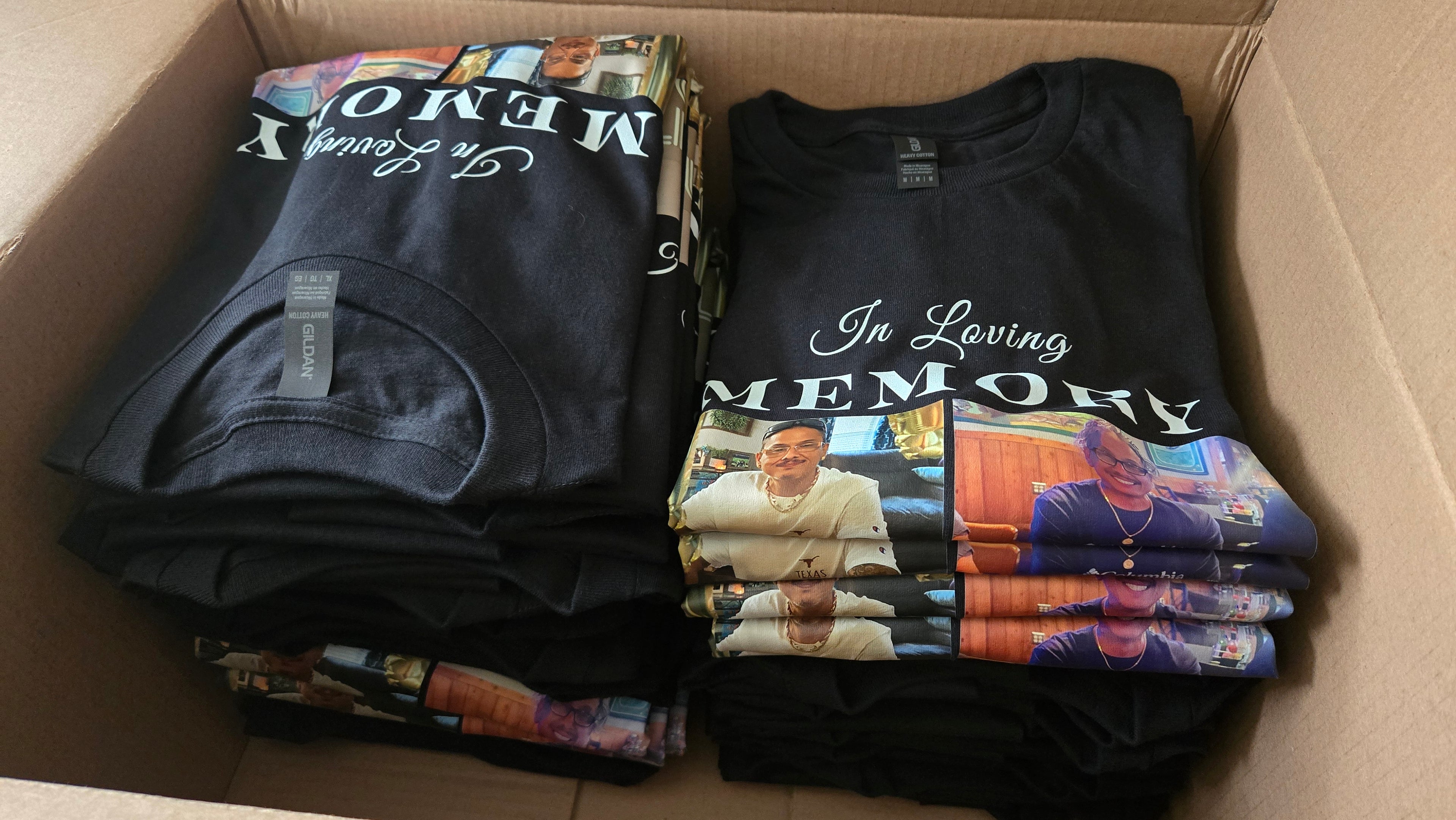 Folded t-shirts with 'In Loving Memory' text and images in a cardboard box.